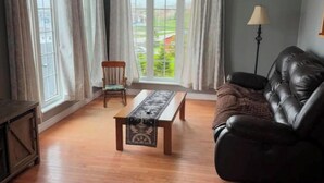 Uma sala de estar com piso de madeira, um sofá de couro preto, uma mesa de centro de madeira, uma poltrona e janelas grandes com cortinas.