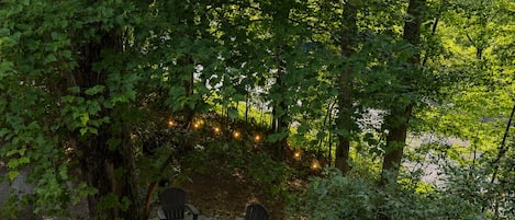 A circular fire pit surrounded by chairs, set in a wooded area with trees and a gravel path.