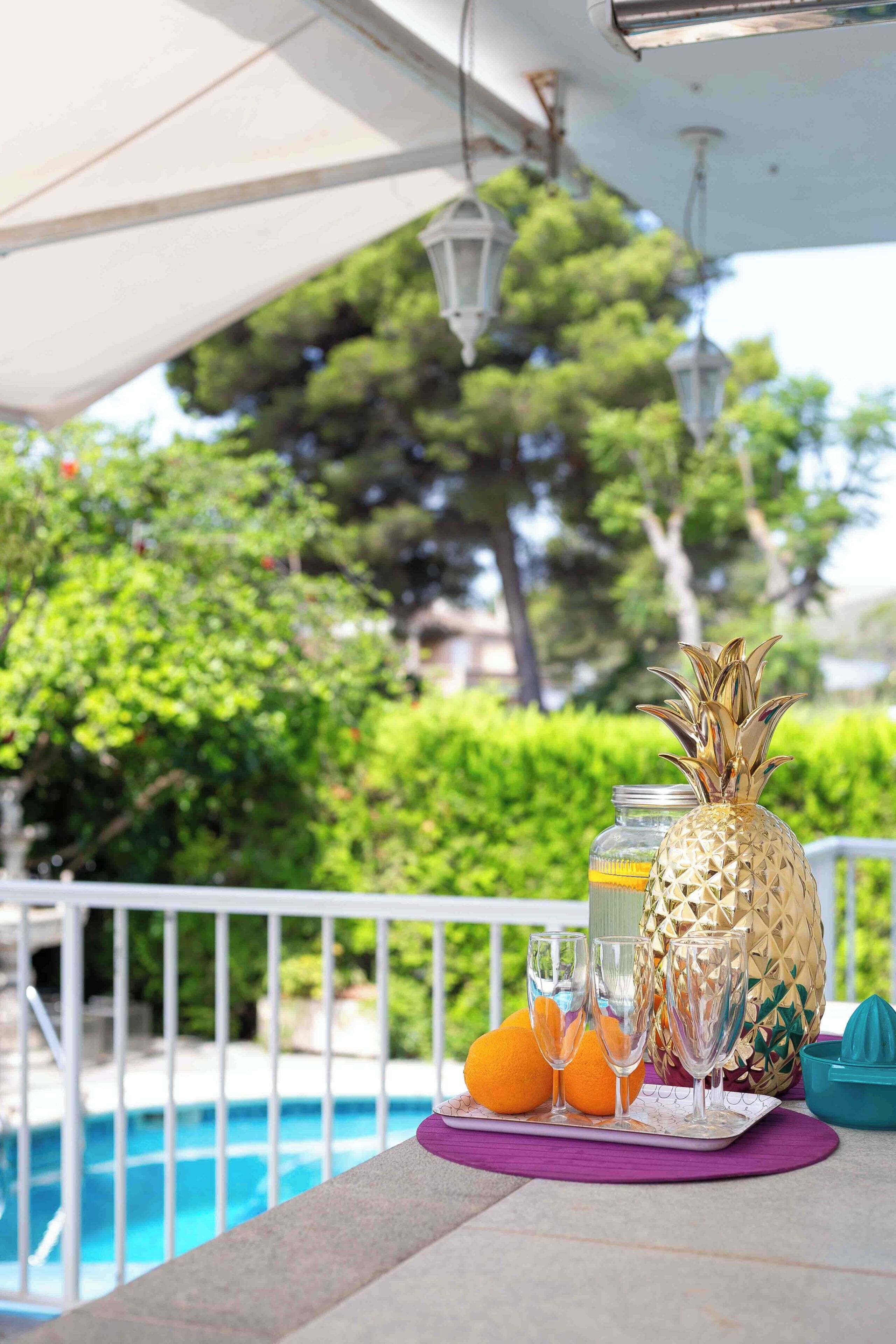 A poolside setting with a pineapple, oranges, and glasses on a table.
