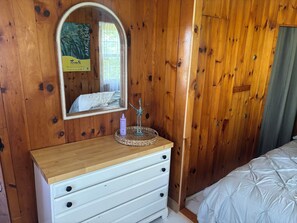 Quarto com painéis de madeira, um penteou branco, um espelho, uma escultura decorativa de vento e uma cama com colcha branca.