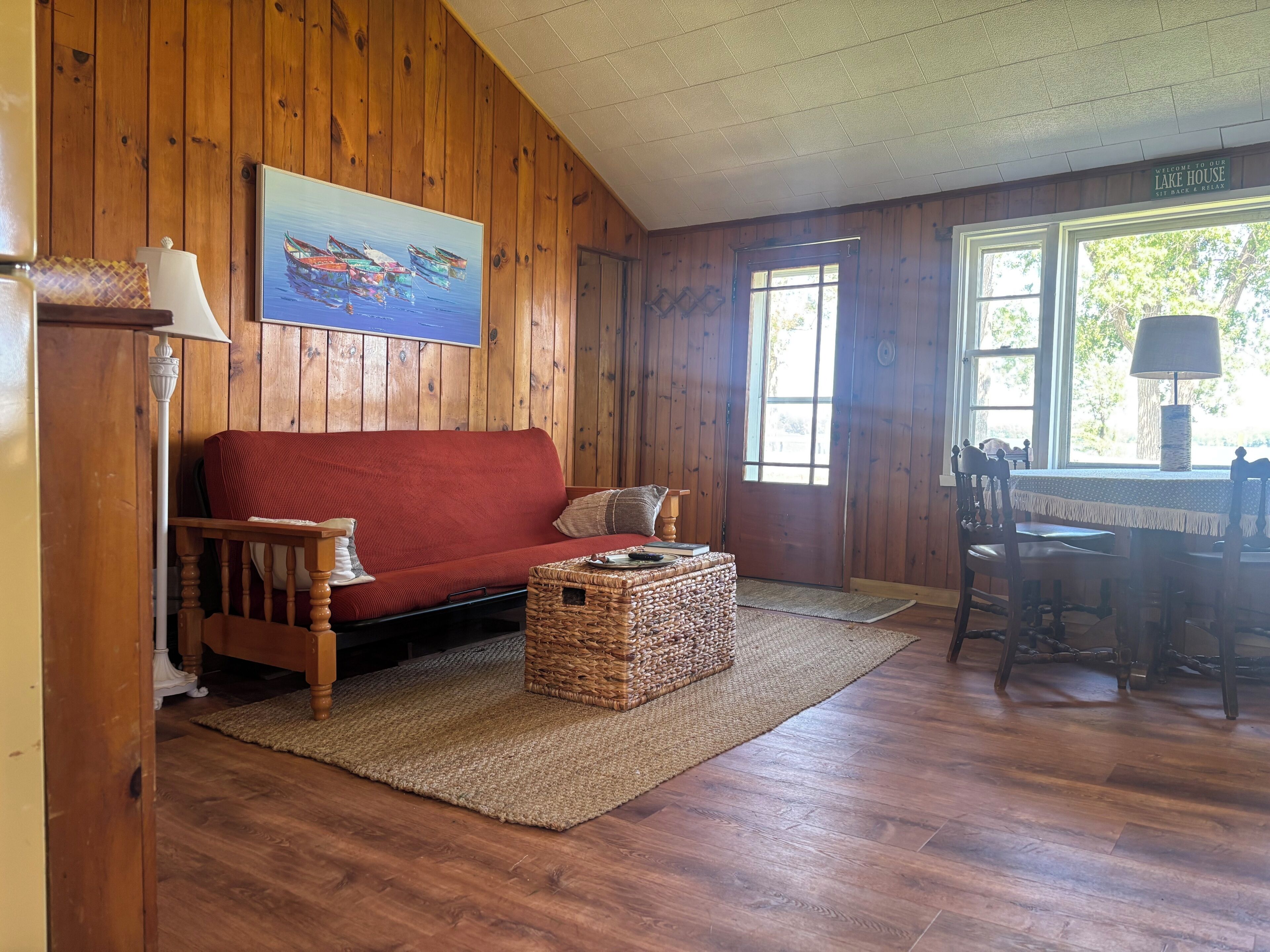 Un intérieur en bois avec un canapé rouge, un panier en osier et un tableau représentant des bateaux.