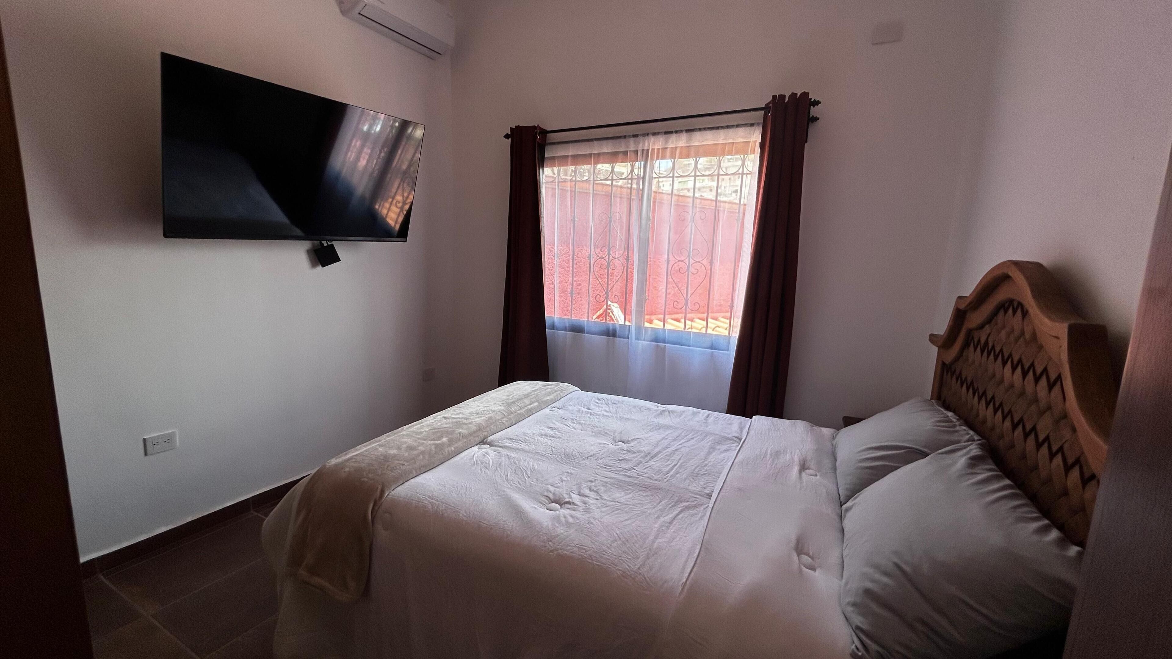 Une chambre à coucher avec un lit, un téléviseur fixé au mur, une fenêtre avec des rideaux et une vue sur l’extérieur.
