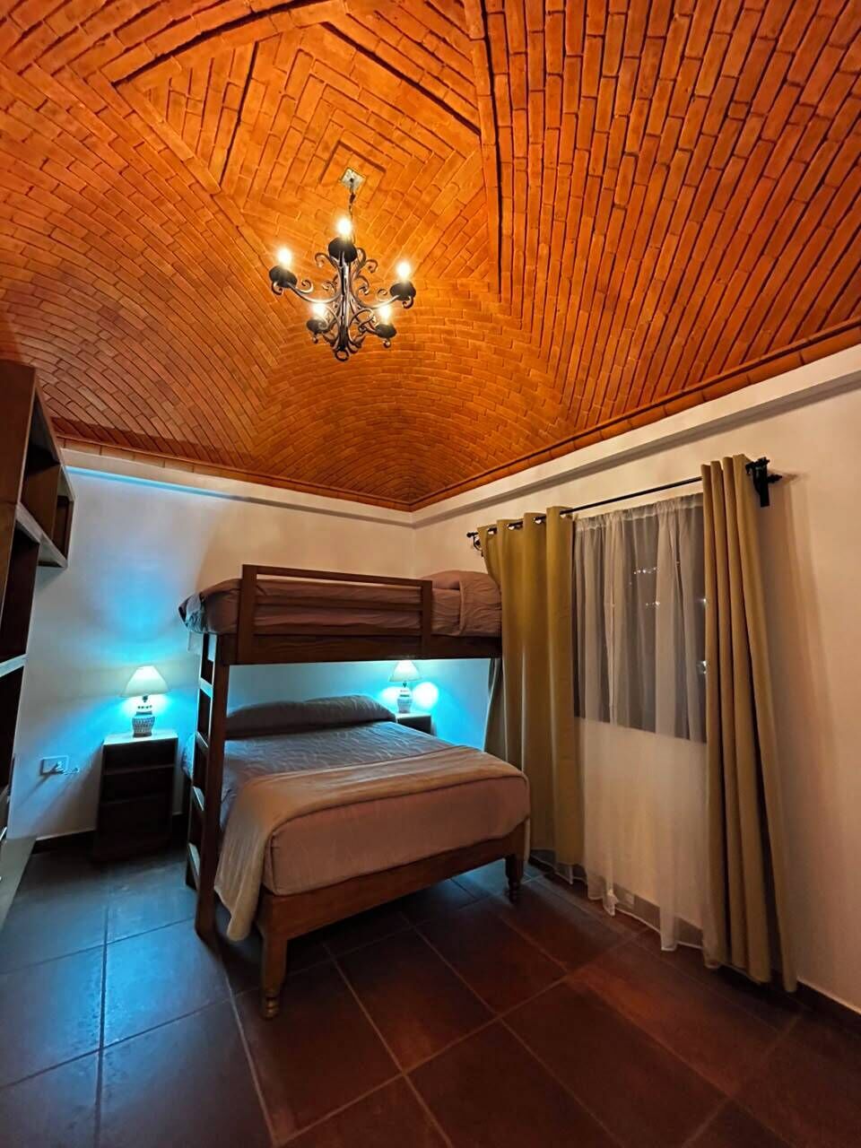 A room with a bunk bed, a chandelier, and a window with curtains.