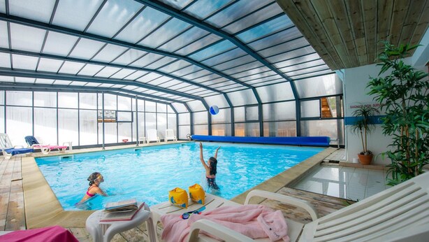 An indoor swimming pool with a glass ceiling, lounge chairs, and a poolside table. - Goelia - Le Village Gaulois (Saint-François-Longchamp)