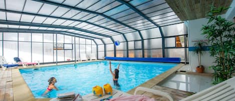 An indoor swimming pool with a glass ceiling, lounge chairs, and a poolside table.