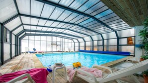 An indoor swimming pool with a glass ceiling, lounge chairs, and a wooden deck.