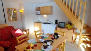 A cozy dining area with a wooden table set for two, a sofa with red cushions, a small kitchen with a microwave and a TV, and a wooden staircase. - Residence Goelia Les Flocons D'argent (Aussois)