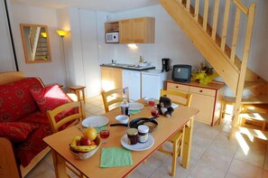 A cozy dining area with a wooden table set for two, a sofa with red cushions, a small kitchen with a microwave and a TV, and a wooden staircase. - Residence Goelia Les Flocons D'argent (Aussois)