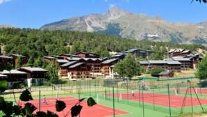 Un complejo turístico de montaña con varias construcciones de estilo chalet, una cancha de tenis y un bosque de fondo.