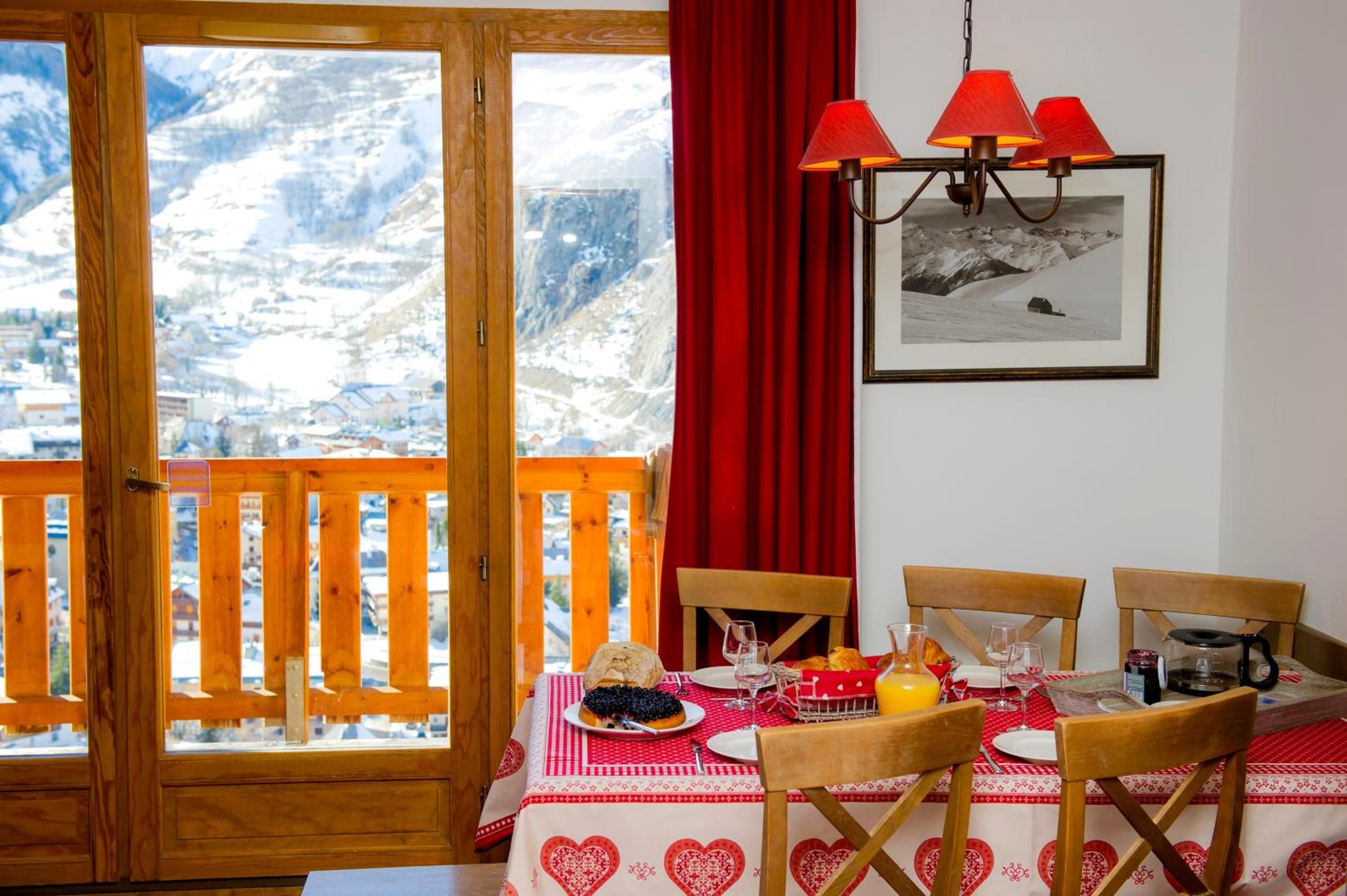 Un coin repas avec une table dressée pour deux personnes, une vue sur la montagne à travers une porte en bois et un rideau rouge.
