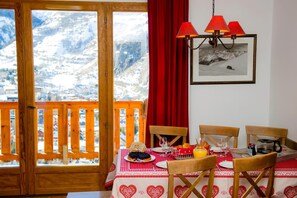 Un coin repas avec une table dressée pour deux personnes, une vue sur la montagne à travers une porte en bois et un rideau rouge.