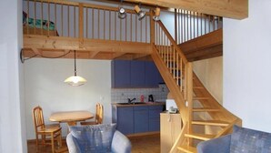 Ett vardagsrum i loftstil med en trätrappa, ett matbord, två blå fåtöljer och ett kök i bakgrunden.