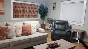 Una sala de estar con un sofá, una mesa de centro, un sillón de cuero negro, una mesita con un teclado y un ventanal con persianas.