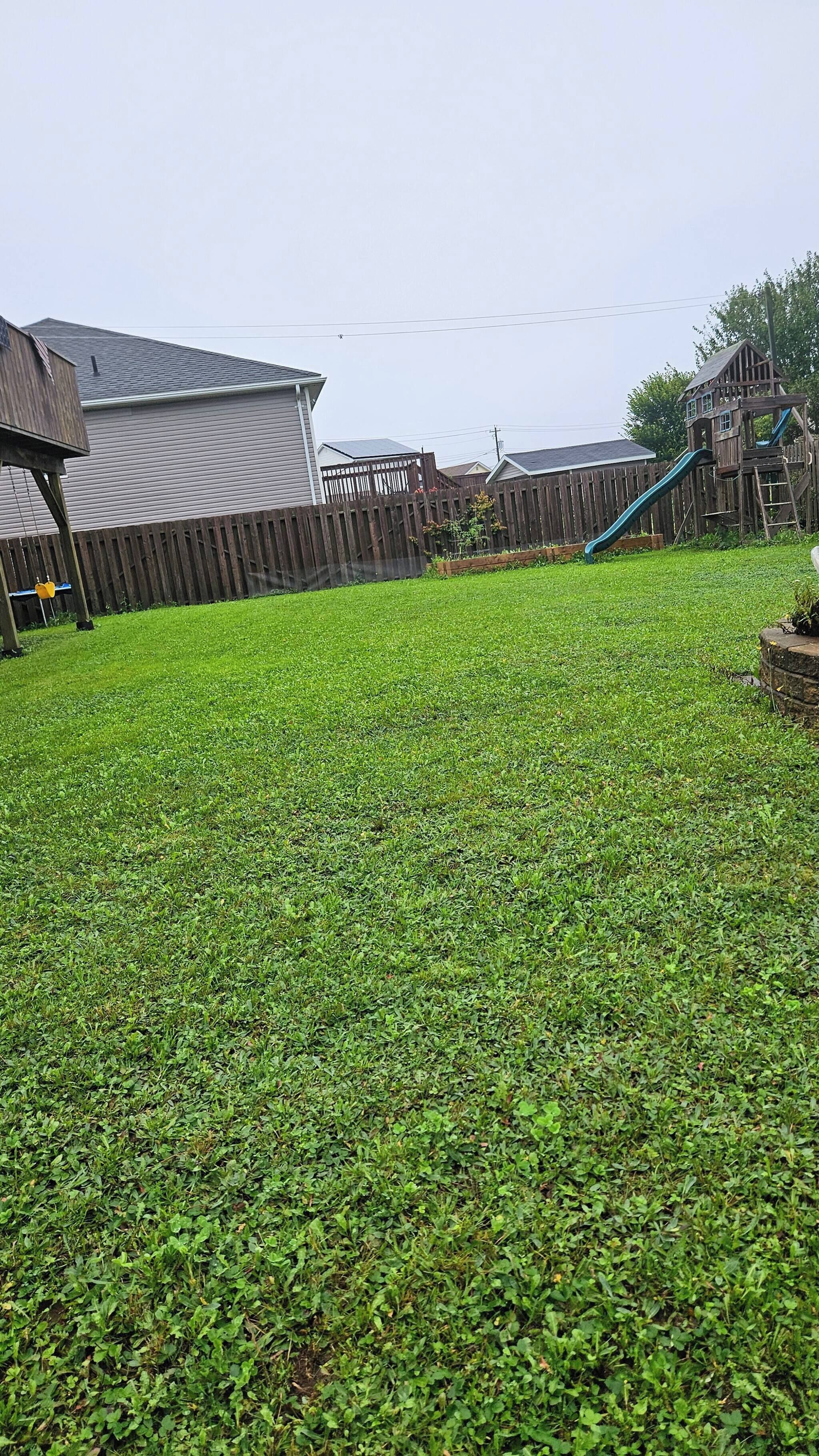Ein Garten mit einer Holz-Spielanlage, einem eingezäunten Bereich und einem Haus im Hintergrund.