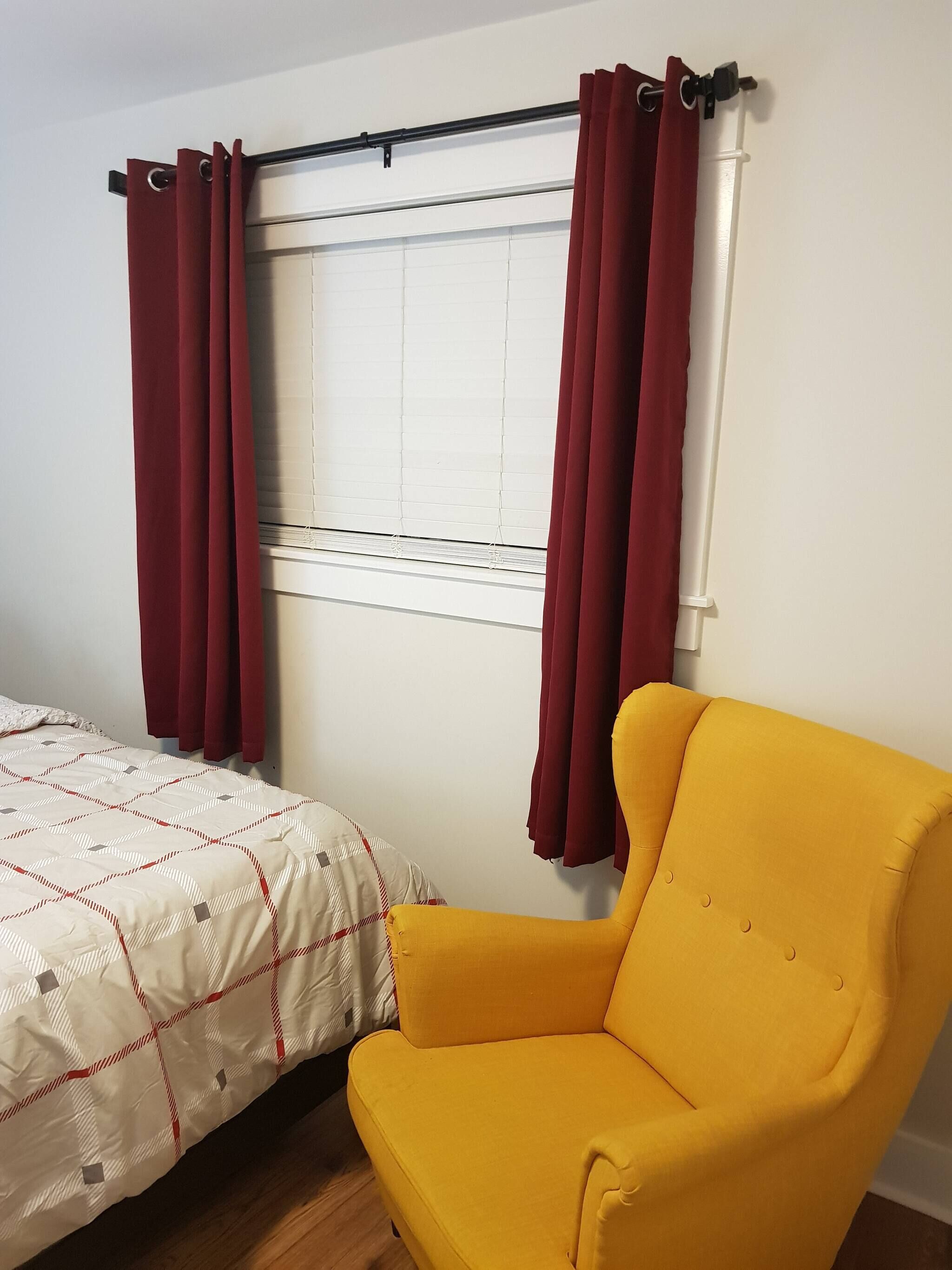 A room with a bed, a yellow armchair, a window with blinds, and red curtains.