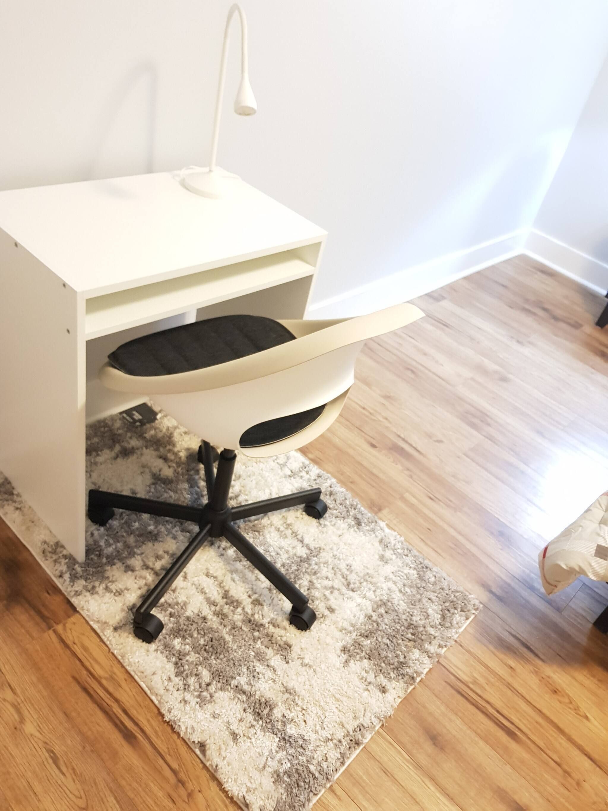 A white desk with a black chair, a small lamp, and a shaggy rug on a wooden floor.