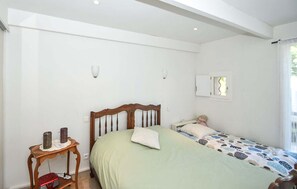 A bedroom with a bed, a nightstand, a small table, a window, and a wall-mounted light fixture.