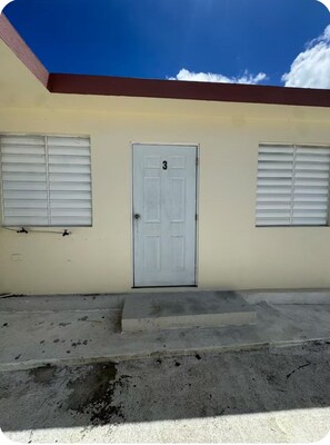 A building with a single door marked with the number 3, two windows with shutters, and a concrete ground. - JG Residences Apartments 3 (san juan)