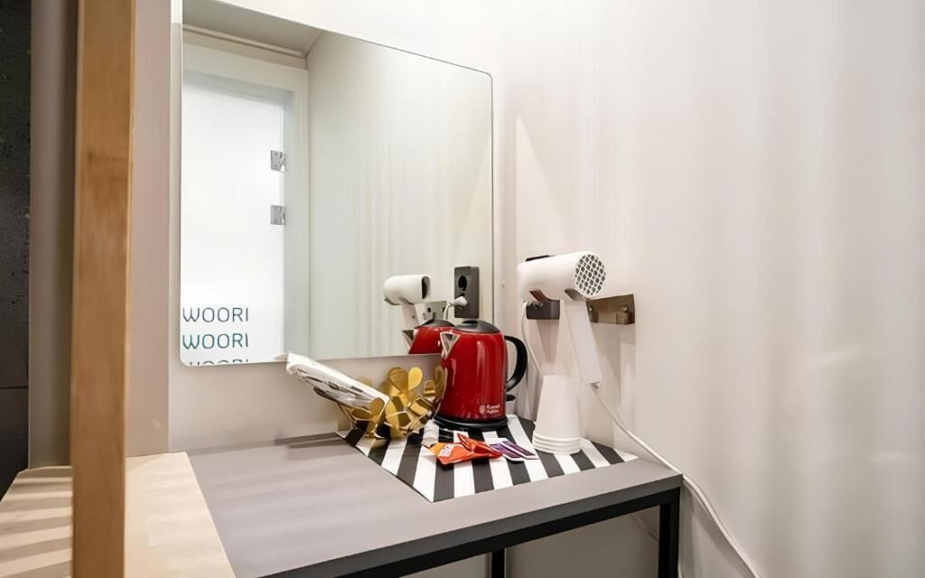 Une salle de bain moderne avec un miroir, une bouilloire rouge, un sèche-cheveux et une petite table surmontée d’un set de table à rayures.