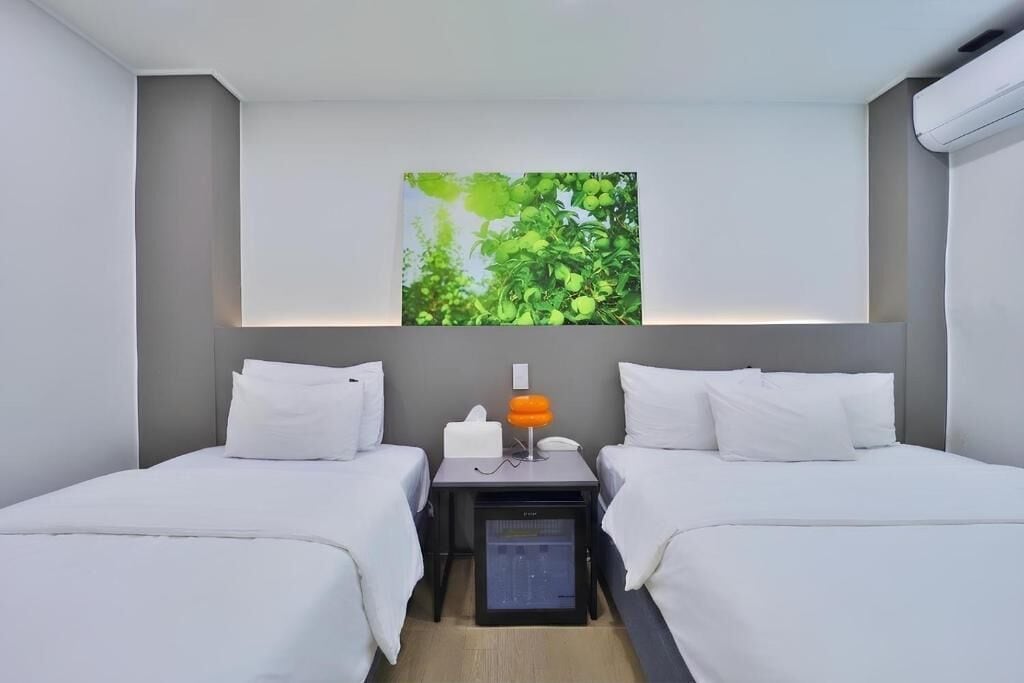 A hotel room with two beds, a nightstand with a lamp, a small refrigerator, and a wall-mounted artwork.