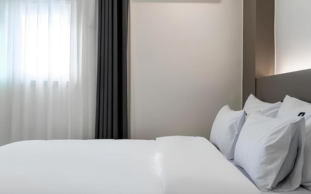 A hotel room with a bed, white linens, a window with sheer curtains, and a modern headboard.