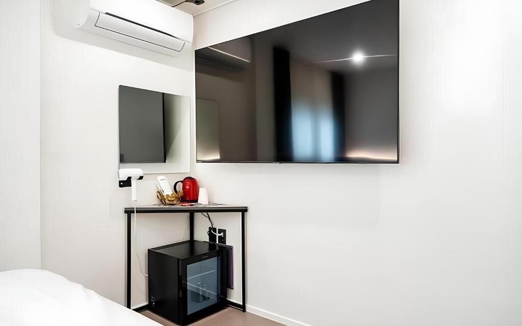 A modern hotel room with a large flat-screen TV mounted on the wall, a small black desk with a kettle, and a built-in mini-fridge.