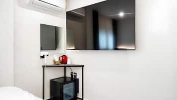 A modern hotel room with a large flat-screen TV mounted on the wall, a small black desk with a kettle, and a built-in mini-fridge.