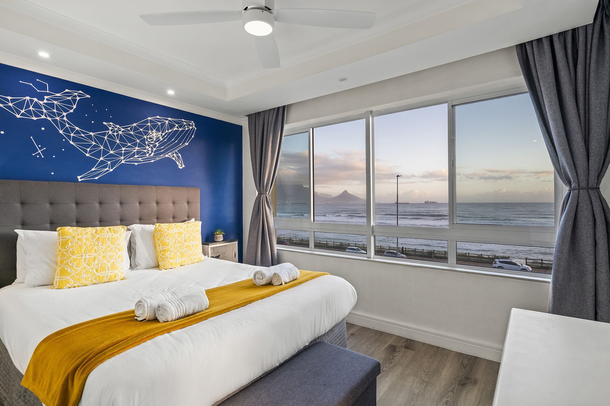 A bedroom with a large bed, a view of the ocean, a geometric wall art, and a ceiling fan.