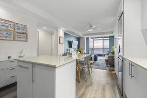 A modern kitchen with a central island, a dining area with chairs, a sofa, and a view of the outdoors. - Ocean View C303 - Two Bedroom Apartment, Sleeps 4 (Cape Town)