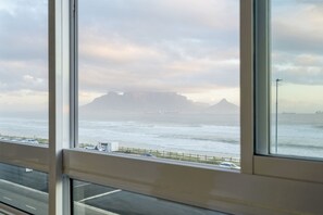 A coastal view through a window, including a road, a beach, and distant mountains. - Ocean View C303 - Two Bedroom Apartment, Sleeps 4 (Cape Town)