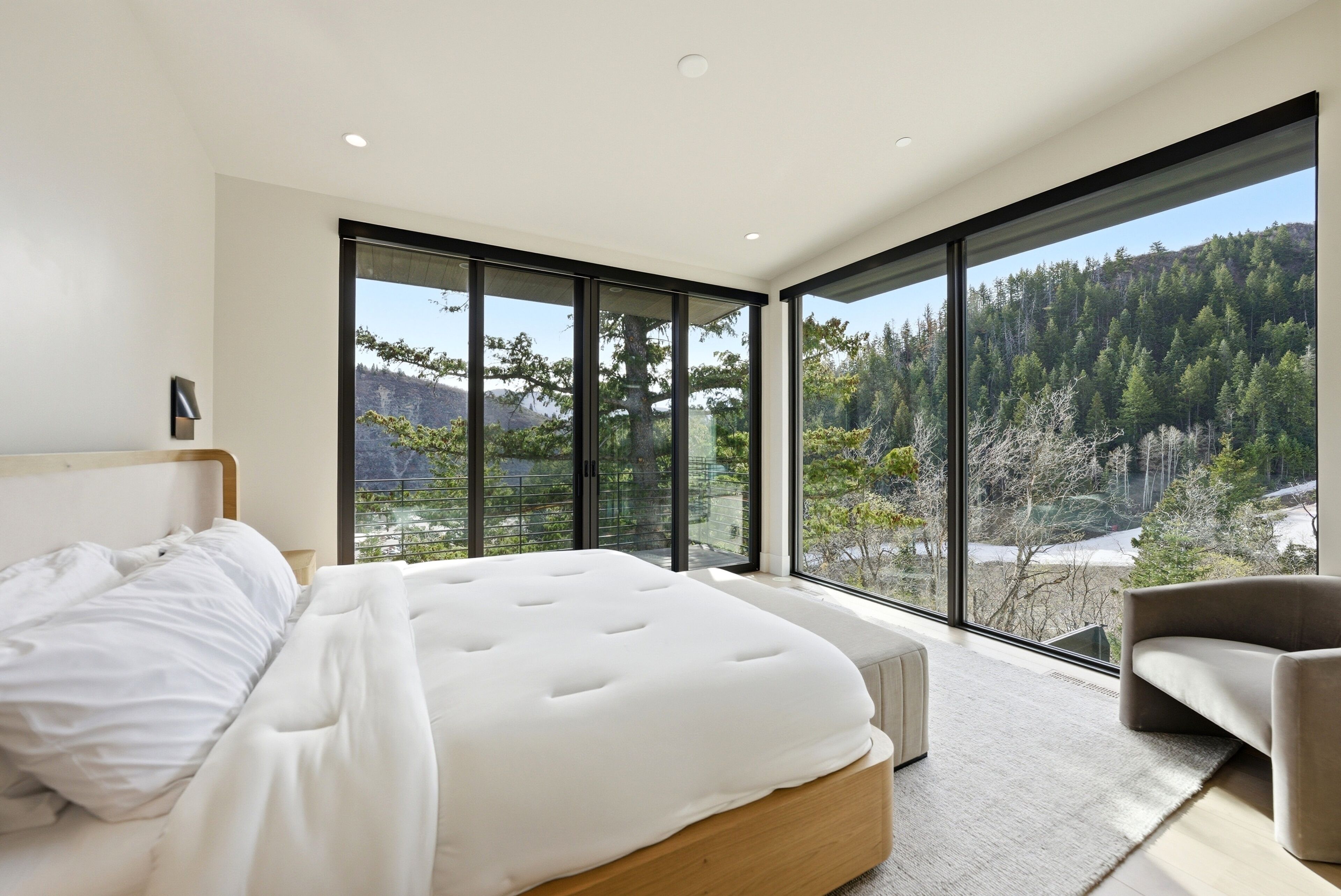 A bedroom with a large bed, a sofa, and a view of a forest through floor-to-ceiling windows.