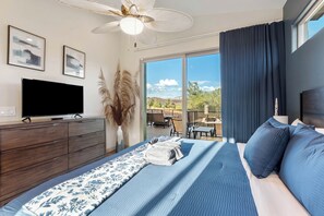 Ein Schlafzimmer mit Bett, Fernseher, Deckenventilator, einem Fenster mit Vorhängen und Blick ins Freie.