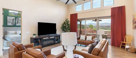 A spacious living room with a flat-screen TV, leather sofas, a wooden coffee table, and a large window offering a view of the outdoors.