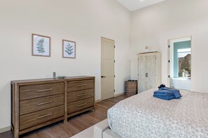 A bedroom with a wooden dresser, a bed with a floral bedspread, a door, and two framed leaves on the wall. - New Luxury Home – Special Introductory Pricing for a Limited Time. (Sedona)