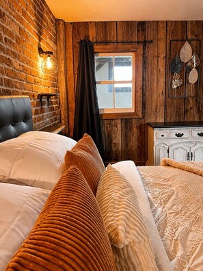 A room with a brick wall, a window with curtains, a bed with multiple pillows, and a dresser with drawers.