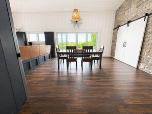 A spacious room with a dining table and chairs, a kitchen area, and a large window offering a view of greenery.