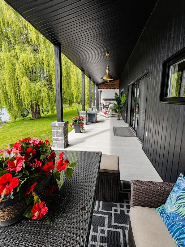 A covered outdoor patio with wicker furniture, a stone planter with red flowers, and a view of a tree and building in the background.