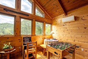 Ein gemütliches Zimmer mit Holzwänden, großen Fenstern, einem Kicker, einem Pac-Man-Spiel und einem Tisch mit Büchern und einer Lampe.
