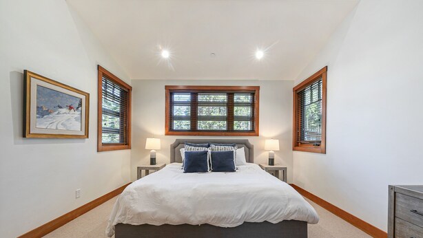 A bedroom with a large bed, two bedside tables with lamps, a window with blinds, and a framed picture on the wall.