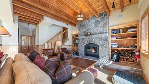 Ein gemütliches Wohnzimmer mit Kamin aus Stein, Holzbalkendecke, einer Treppe und einem Bücherregal voller Bücher.