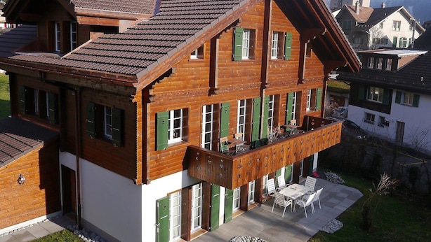 Een meerlaags houten gebouw met een balkon en een terras met zitplaatsen buiten, met een bergachtig landschap op de achtergrond.