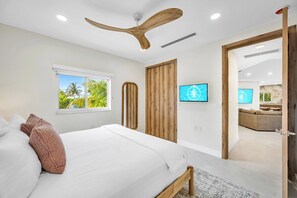 A modern bedroom with a bed, a ceiling fan, a TV, and a view of a living area.