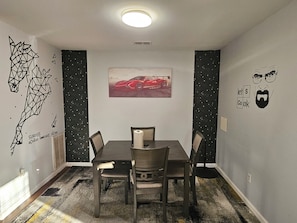 Un espace repas comprenant une table, des chaises, un luminaire au plafond et des éléments de décoration murale, notamment une affiche de voiture et des œuvres d’art géométriques.
