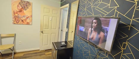 Ein Zimmer mit an der Wand befestigtem Fernseher, einer Tür, einem Stuhl und einem kleinen Tisch mit Fernbedienung.