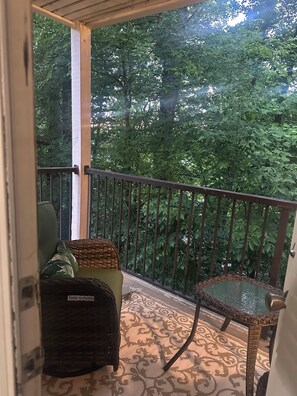 A balcony with a wicker chair, a small table, and a view of dense foliage. - Vinings Luxe Escape Luxury 2 Bd In Atlanta (Atlanta)