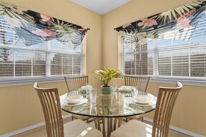 Un coin repas comprenant une table ronde en verre, six chaises, un vase avec des fleurs et deux fenêtres équipées de stores.