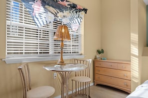 Ein Zimmer mit einem runden Glastisch, zwei Stühlen, einer Holzkommode und einem Fenster mit Jalousien und einem blumigen Vorhang.