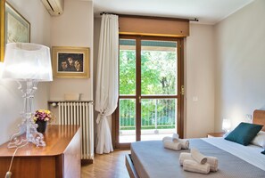 Een hotelkamer met een bed, een bureau met lamp, een radiator, een raam met gordijnen en een ingelijst schilderij aan de muur.