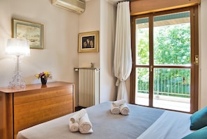 Habitación de hotel con cama, cómoda de madera, lámpara, radiador y ventana con vistas a zonas verdes.