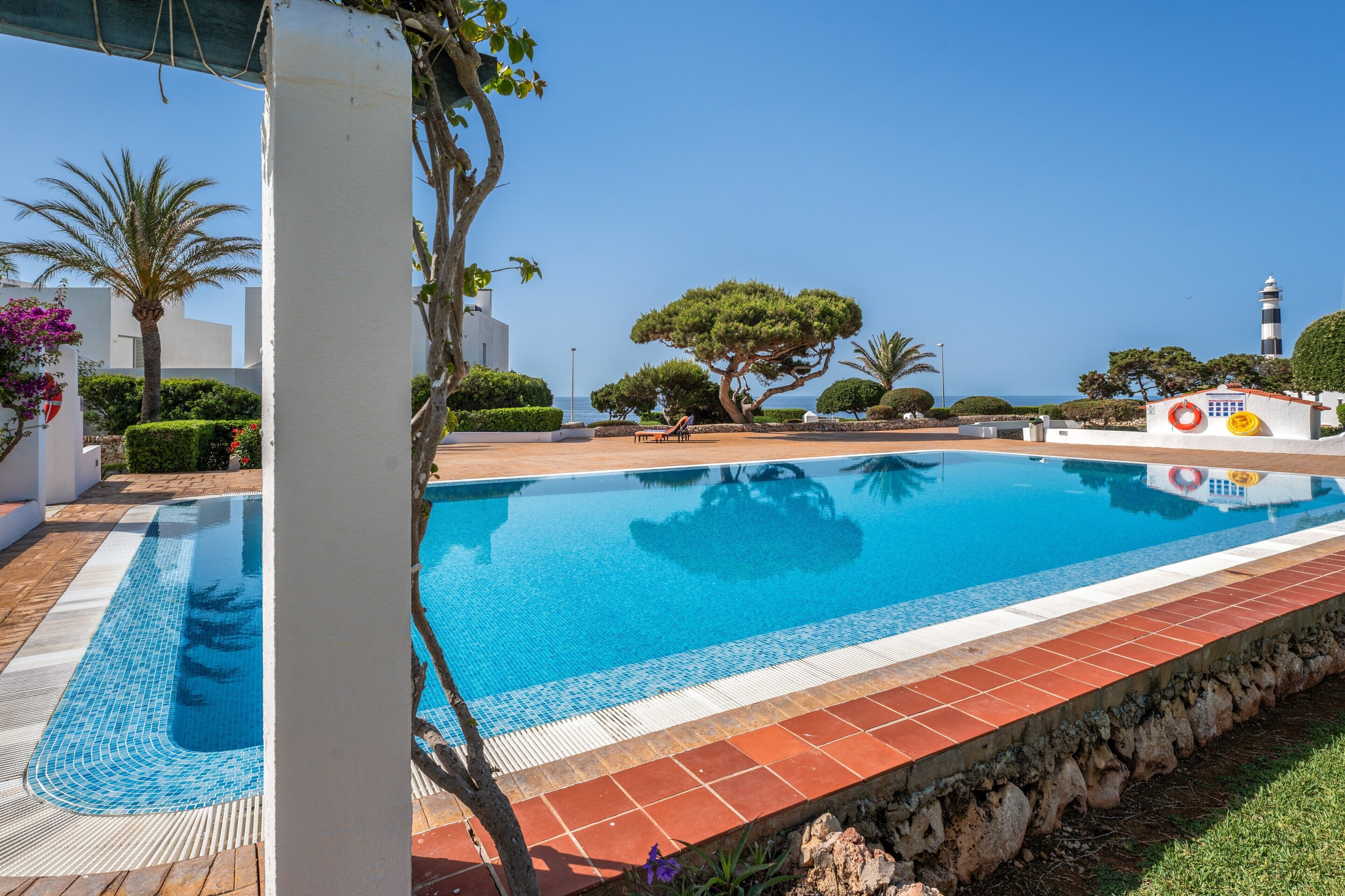 A swimming pool with a tiled edge, surrounded by greenery and a clear sky.